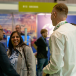 Three people stand and converse in a busy exhibition area, with booths and other attendees visible in the background.